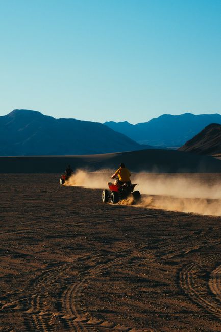 Two people riding ATVs in a desert, kicking up dust