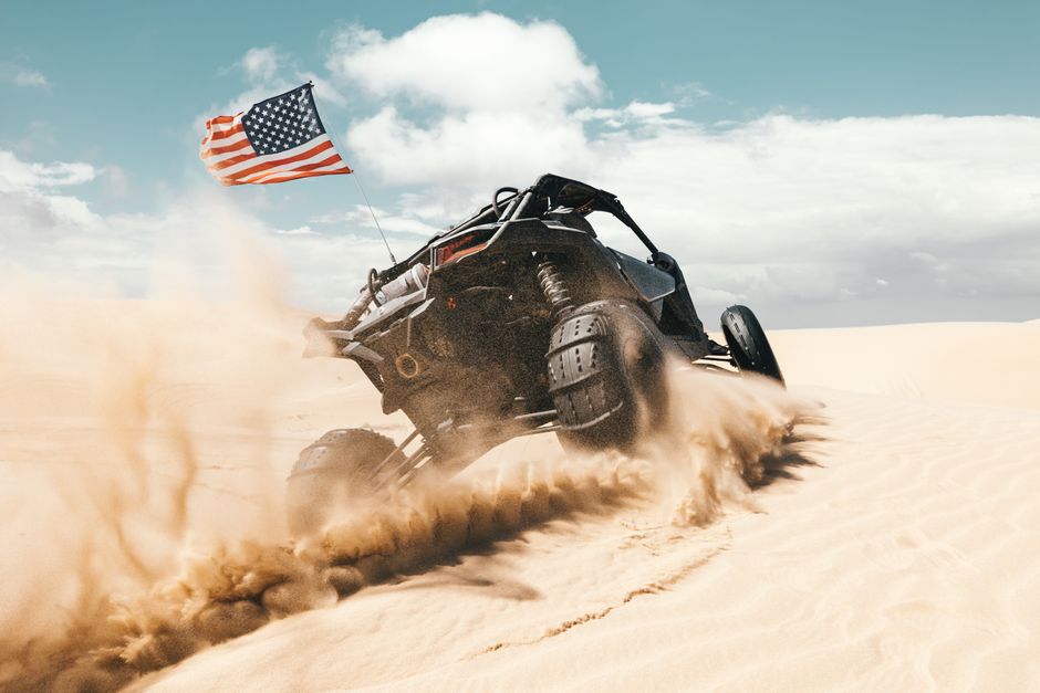 A black desert buggy kicking up sand, with an American flag flying