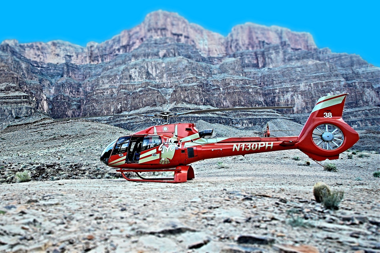 A red helicopter with white stripes and the text 'N130PH' on its side, parked on a rocky surface with a large canyon in the background.