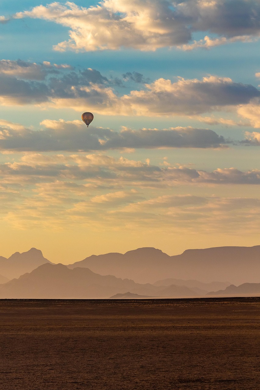 A hot air balloon floats in a cloudy sky above a desert landscape and mountains at sunrise or sunset.