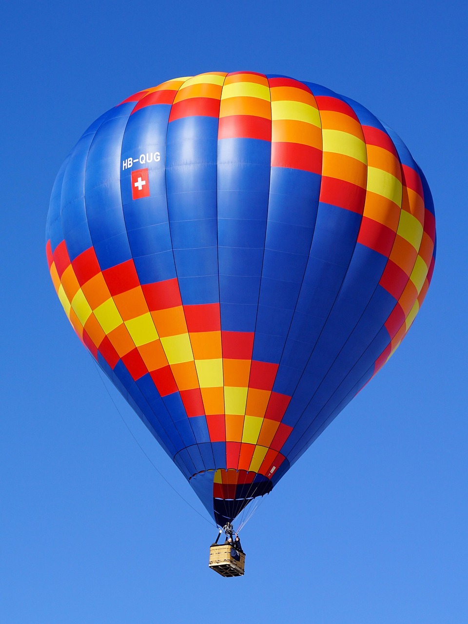 A hot air balloon with a blue, red, orange, and yellow striped envelope flies against a clear blue sky.