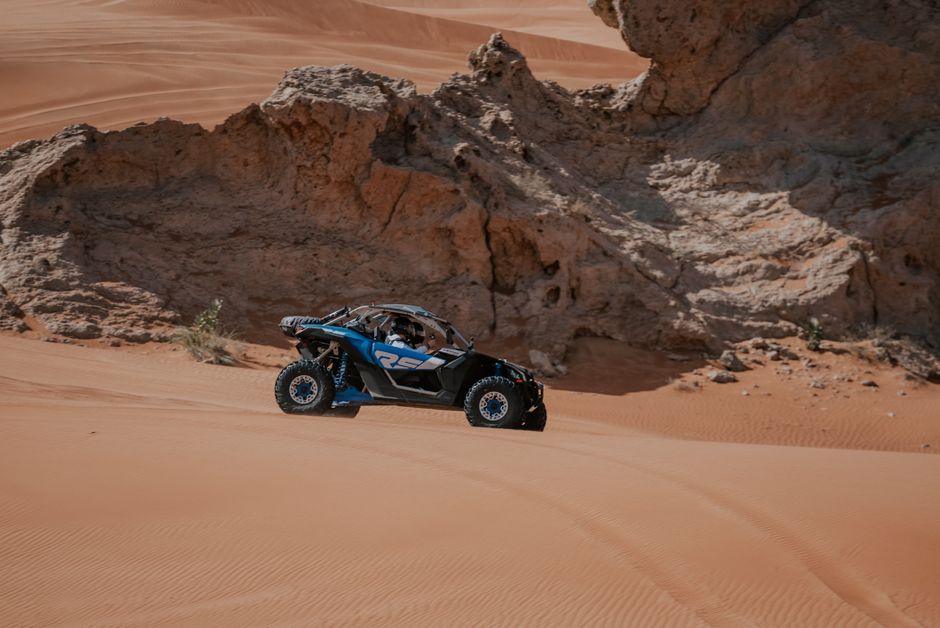 A blue and black UTV driving on sand dunes with rocky outcroppings in the background