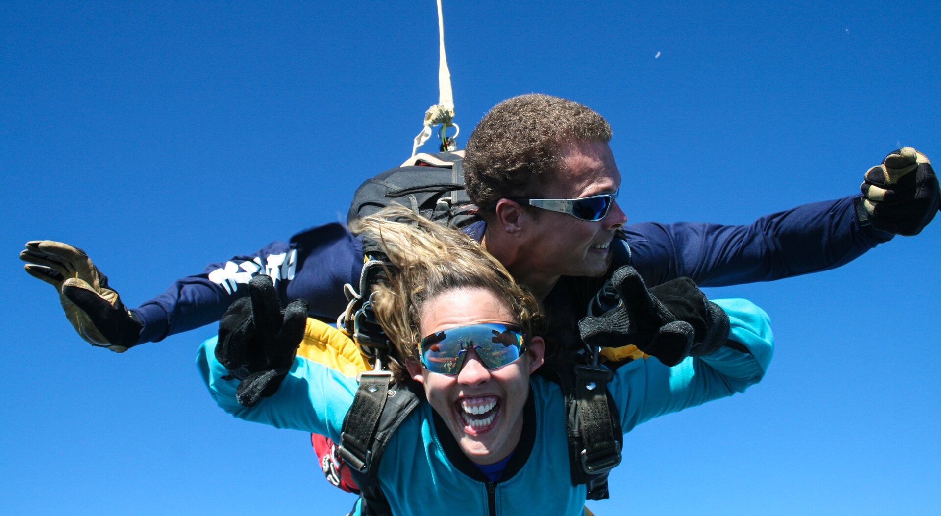 Two people in skydiving gear, one attached to the other, against a blue sky