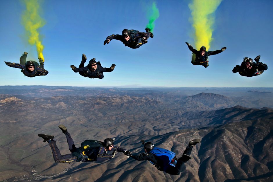 seven skydivers in formation with colored smoke trails over a desert landscape