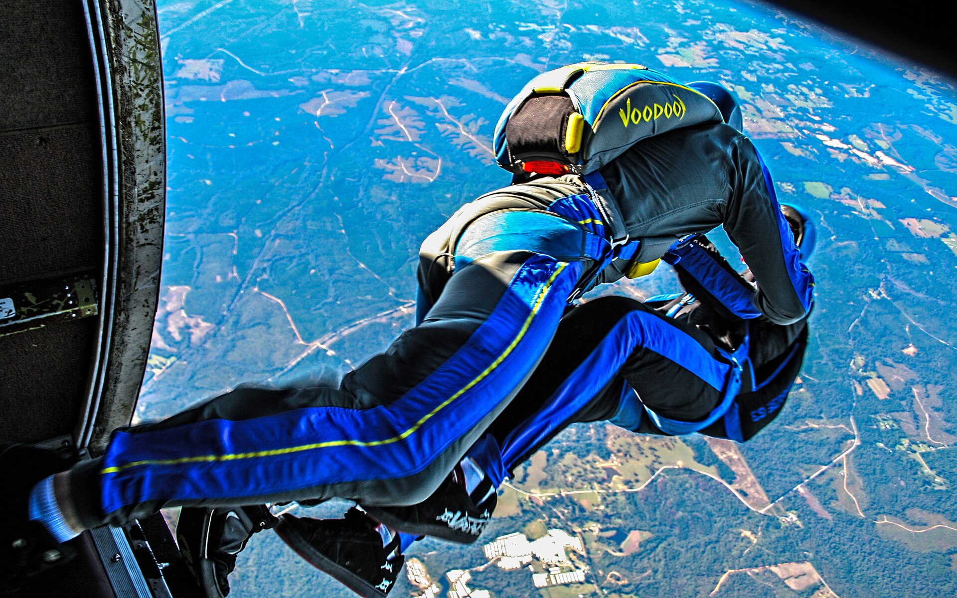 Two skydivers exiting a plane, one wearing a blue and black jumpsuit with 'Voodoo' on the helmet
