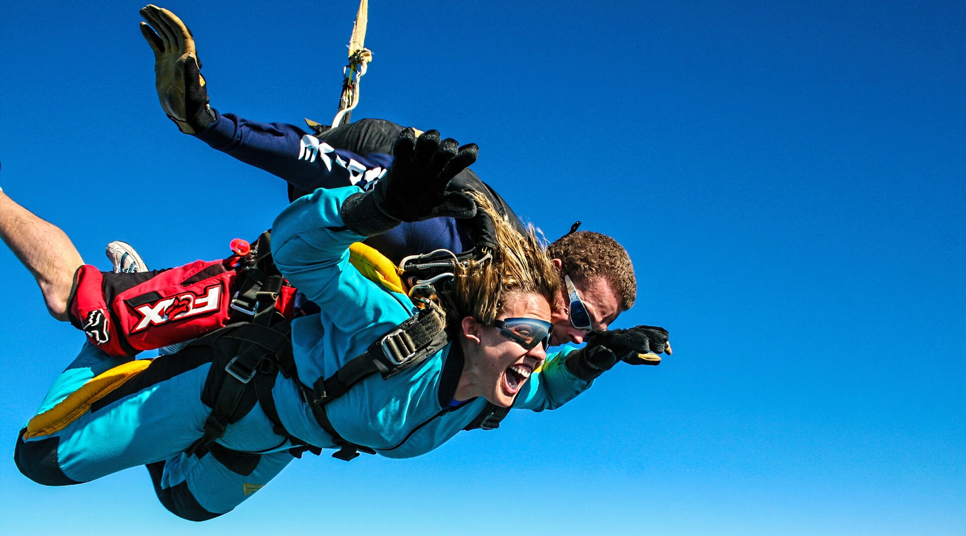 Two people skydiving, one attached to the other's back, against a clear blue sky