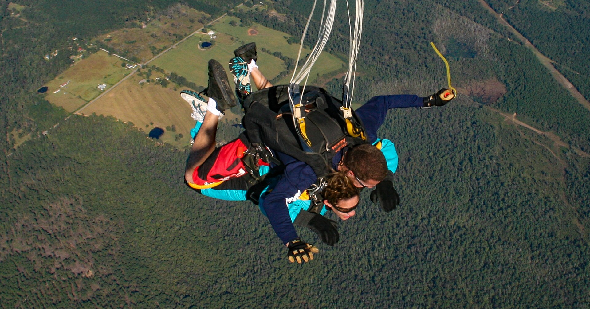 Two people skydiving, one attached to the other, over a forested landscape