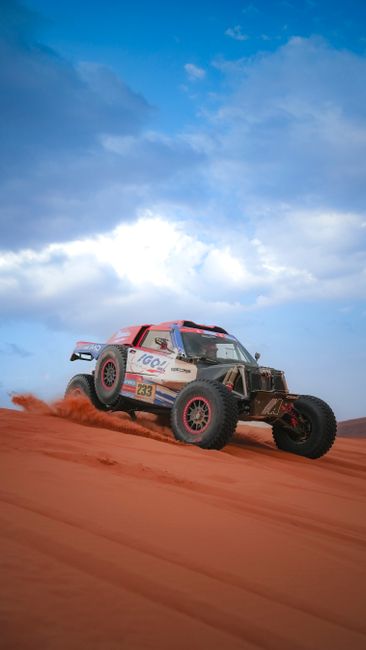A white and black off-road vehicle with large tires kicking up red sand on a desert track