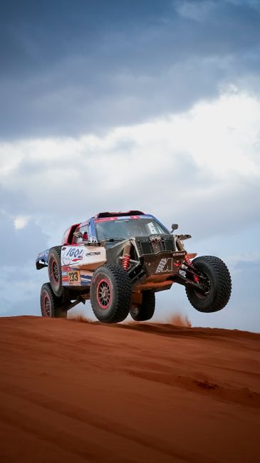 A white, red, and blue off-road vehicle airborne on a red sand dune