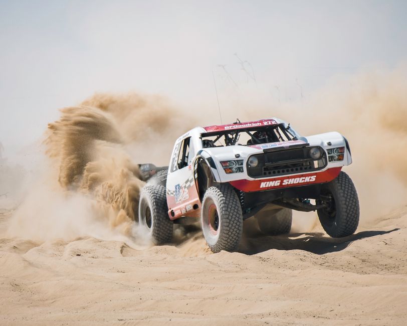 A white and red off-road truck driving on sand, kicking up dust